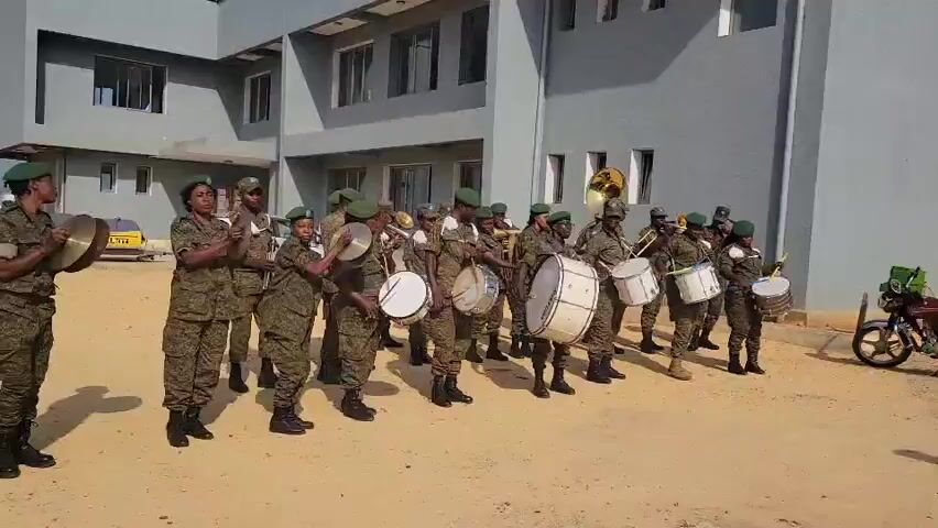 The video shows a military band, composed of approximately 15 individuals in camouflage uniforms and green berets, standing in formation in front of a light gray building. The band members are playing various instruments, including drums and brass instruments. Some are holding cymbals. The ground appears to be dirt or sand. The building has multiple windows and appears to be modern. The lighting suggests it is daytime. There is a yellow vehicle visible in the background to the left of the building. To the right, part of a motorcycle or similar vehicle is visible. The band members are facing forward, and some appear to be actively playing their instruments.