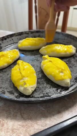 The video shows a close-up shot of a dark, round metal baking pan containing five crescent-shaped pastries. The pastries are raw dough, and each one is coated with a bright yellow liquid, presumably an egg wash. A wooden-handled brush is visible at the top right corner, in the process of applying the yellow coating to one of the pastries. The pan is sitting on a surface that appears to be a countertop, with a patterned tablecloth or mat underneath. In the background, a portion of a wooden chair and a wall with a decorative element are visible. The lighting is soft, suggesting an indoor setting. The overall scene depicts the preparation of pastries before baking.