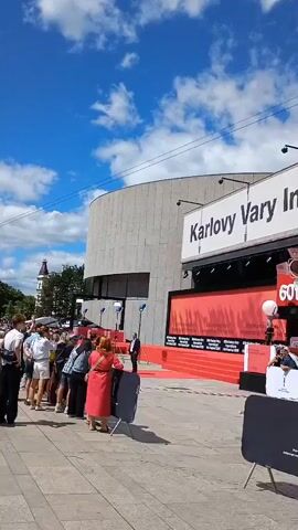 A video begins by showing a sign that reads "Karlovy Vary International Film Festival" above the entrance to a building. Below the sign is a red carpet leading into the building. Several people are standing on the red carpet, and some are waiting in line. There are also some barriers and tents set up in front of the building. The video pans to the left, showing more of the building's exterior, which is made of concrete. The sky is blue with some clouds. The video continues to pan across the scene, showing more people and tents. It ends by focusing on the entrance to the building, where a few people are walking inside.