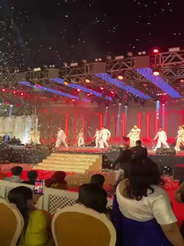 The video shows a group of dancers performing on a stage in front of an audience. The stage is decorated with lights and confetti, and the dancers are wearing white outfits. The audience is seated in chairs and watching the performance. The scene is brightly lit and appears to be part of a larger event.