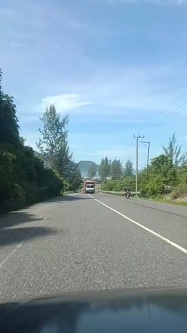 The video shows a view from a vehicle driving on a paved road. The road has lane markings and a guardrail on the right side. Trees and lush green vegetation line the left side of the road, and more trees are visible on the right. In the distance, there is a body of water, likely the ocean, and a small island or landmass. The sky is blue with some scattered clouds. A white truck with a red top is driving ahead on the road. Further ahead, a motorcycle with a rider is also visible on the right side of the road. The scene appears to be a coastal road in a tropical or subtropical region. The video is short and appears to be a single continuous shot.