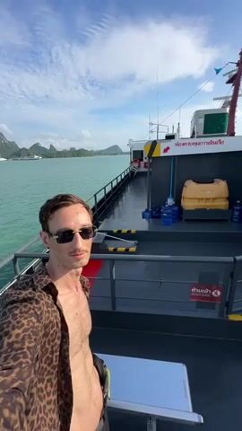 The video opens with a man with short brown hair and a mustache wearing sunglasses and an open leopard-print shirt standing on the deck of a ferry. He is holding a camera and taking a selfie. The ferry is moving on a turquoise body of water. In the background, there are several lush green islands with jagged mountain peaks. The sky is blue with scattered white clouds. The deck of the ferry is dark gray and has a metal railing. On the right side of the deck, there are several large yellow containers and blue water jugs. There is also a red fire extinguisher mounted on the railing. The ferry has some signage in a foreign language. The man is looking at the camera with a slight smile. The camera pans slightly to the left, showing more of the water and the distant islands. The video continues with similar shots of the man and the surrounding scenery.