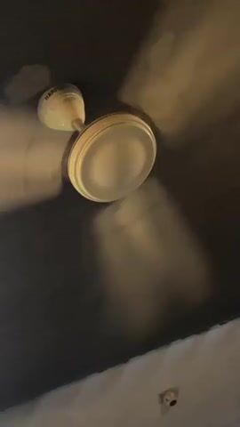 The video starts with an upward shot of a round, diffused ceiling light fixture and a small, light-colored sensor on a dark ceiling. It then quickly transitions through blurry shots before settling on an outdoor evening scene. The main subject is a young woman with dark, curly hair, adorned with a beaded headband (possibly shells) and flowers in her hair. She wears multiple rings on her fingers and a dark garment with a purple floral pattern, looking at the camera with a slight smirk. She appears to be seated at a table or counter, with a green railing visible in the foreground. The background shows an outdoor area illuminated by warm string lights, green foliage, and other indistinct figures, indicating a social gathering at night in Korgao, India. No text is visible.