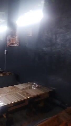 This is a wide shot of an indoor restaurant with dark walls and wooden tables and benches. The lighting is bright and overhead. The camera pans across the room, showing several tables set with glasses and a few framed pictures on the wall. The room appears to be empty of customers.