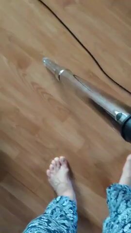 A low-angle, first-person perspective video shows a person's bare feet and lower legs, clad in blue and white patterned pants, standing on a light brown wood-patterned floor. A dark-colored vacuum cleaner hose with a metal extension tube and a nozzle is visible in the upper part of the frame, moving back and forth across the floor, suggesting the person is vacuuming. A black power cord is also visible on the floor to the right.