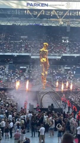 A wide shot shows a concert stage setup in a stadium filled with a large audience. In the center of the stage stands a very tall, golden, humanoid statue. The statue is positioned above a circular, metallic structure that resembles a tunnel or ring. Smoke and pyrotechnic effects, including bursts of orange flames, are visible around the base of the statue and on the stage. The crowd is densely packed, facing the stage. In the background, stadium seating is filled with spectators. Overhead, a large banner displays the word "PARKEN" and "CONNECTED BY 3." The lighting suggests an evening event.