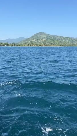 The camera is positioned on a boat, capturing a view of the ocean. The water is a deep blue, with small waves creating a textured surface and whitecaps visible near the boat. In the distance, an island is visible, dominated by a large, rounded green hill covered in trees and vegetation. A shoreline with palm trees and other foliage stretches along the base of the hill. Clear blue sky with no clouds above the water and island.