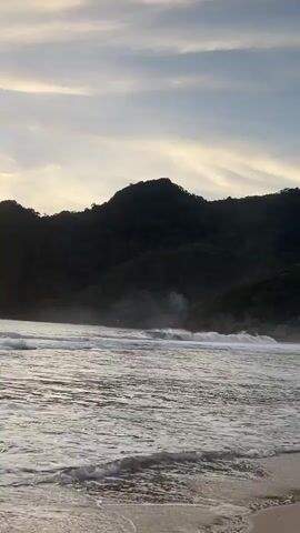 The video shows a tranquil beach scene at dusk. Gentle waves lap the sandy shore, creating a soothing rhythm. Lush green hills form a dramatic backdrop to the serene ocean. The sky above is a canvas of soft clouds, tinged with the warm hues of the setting sun. A few wisps of smoke are visible rising from the treeline on the hills, adding a touch of mystery to the otherwise peaceful panorama. The overall atmosphere is one of quiet beauty and natural splendor.
