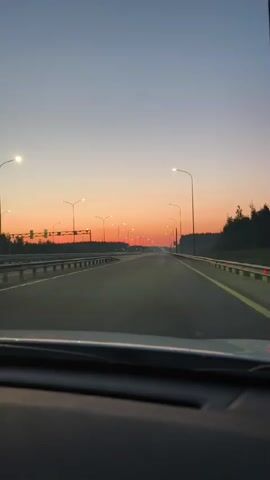 The video shows a first-person view from inside a car driving on a highway at dusk. The sky transitions from a deep blue at the top to a vibrant orange and pink gradient near the horizon. Numerous streetlights line the sides of the highway, their lights illuminated. The road ahead is empty, with multiple lanes clearly marked. On either side of the highway, there are rows of trees. The dashboard of the car is visible in the foreground, indicating the perspective of the driver.