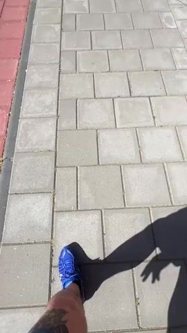 The video shows a first-person view of someone walking on a sidewalk. The person is wearing blue sneakers. The sidewalk is made of gray paving stones and is bordered by a red-brick sidewalk. To the right of the sidewalk is a red-brick building with a white door. To the left, there is a grassy area with trees. A black lamppost is visible along the sidewalk. The shadow of the person holding the camera can be seen in the video. The weather appears to be sunny.