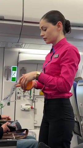 A female flight attendant is standing in the aisle of an airplane, giving instructions to passengers. She is holding a yellow oxygen mask in one hand and a safety instruction card in the other. The flight attendant is wearing a pink button-down shirt with a blue collar, black pants, and a gold watch. Her hair is pulled back in a bun. Passengers are sitting in their seats, with one passenger holding a phone. The emergency exit sign is visible on the wall behind the flight attendant.