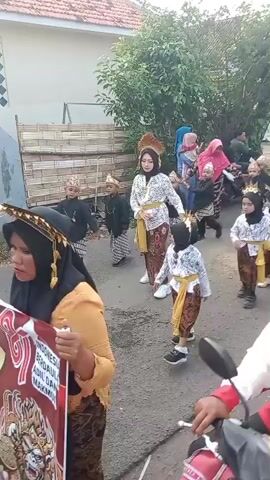 A group of children dressed in traditional Indonesian attire are walking down a street, some carrying banners. They appear to be participating in a parade or cultural event. The children are wearing sarongs, batik shirts, and headpieces. Several adults are also present, some of whom are holding the banners and guiding the children. The environment appears to be a residential area, with houses and trees visible in the background.