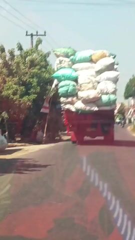 The video shows a large truck overloaded with sacks of cargo. The sacks are stacked very high, exceeding the height of the truck's cab and appearing to be unstable. The truck is driving on a paved road during the daytime. The road appears to be in a rural or semi-rural area, with houses and trees visible on the sides. There are also utility poles and wires in the background. A few motorcycles are seen on the road in the distance. Indonesian flags are displayed on poles along the roadside, suggesting the location may be Indonesia. The truck appears to be moving forward at a moderate speed. The camera is positioned from a vehicle following behind the truck.
