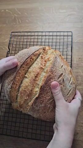 A close-up shot shows two hands holding a round loaf of bread with a golden-brown crust. The bread is resting on a black wire cooling rack, which is placed on a wooden surface. The hands are positioned on either side of the loaf, gently turning it to reveal its textured surface, which is scored with decorative patterns. The lighting is natural, casting soft shadows and highlighting the crust's crisp appearance. The background is a blurred wooden table.