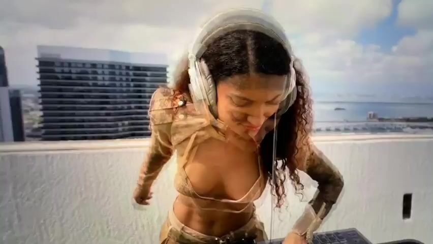 The video shows a woman with long, curly brown hair wearing headphones and a stylish outfit consisting of a long-sleeved top with cutouts and a matching skirt. She is standing on a rooftop overlooking a city skyline with a body of water. In the foreground, a DJ controller is visible, suggesting she is a DJ. A camera on a tripod is also positioned nearby, indicating a possible filming or photography session. The woman appears focused on the DJ controller and the music.

The setting appears to be an urban environment, likely a city, given the presence of tall buildings. The time of day seems to be daytime, with clear skies and visible sunlight. The woman's expression is one of concentration, but she also seems to be enjoying herself, as evidenced by her slight smile and head movements in some frames. The overall mood is energetic and vibrant, hinting at a performance or creative process.

There is no visible text in the video.