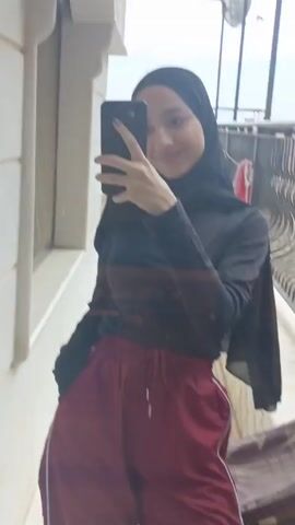 A young woman in a black hijab is taking a selfie with a black smartphone. She's wearing a long-sleeved black shirt and red sweatpants with white stripes down the side. She's on a balcony with a decorative metal railing. A satellite dish and other buildings are visible in the background.