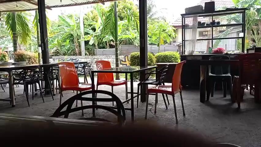 This video shows an outdoor seating area of a restaurant. There are several tables and chairs, some of which are orange plastic and others are black metal. In the background, there are banana trees, a fence, and a building. There are no people visible in the video. The lighting suggests it is daytime.