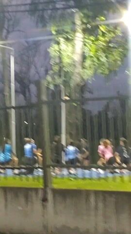 The camera, positioned outdoors, is looking through a fenced perimeter at an outdoor basketball court. There are several people present on the court, though the distance and poor lighting make it difficult to discern the exact number or their actions. Tall light poles illuminate the area with bright, stark light, creating a sharp contrast against the dim and overcast sky above. The ground in the foreground is covered with grass. The focus of the camera seems to shift slightly during the recording, occasionally improving clarity.