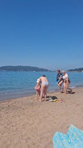 The video shows a beach scene on a sunny day with a clear blue sky. In the foreground, the sandy beach is visible. A body of water, likely the sea, stretches out into the background, with a distant coastline featuring hills and trees. Several people are present. Close to the water's edge, a woman in a bikini is interacting with a small child. Another woman, wearing a t-shirt and shorts, walks away from the water, carrying a blue object. A third woman, also in a t-shirt and shorts, is partially visible on the left side of the frame. There are some colorful objects, possibly children's toys, on the sand. In the distance, a jet ski is visible on the water. The overall atmosphere is one of a pleasant day at the beach.