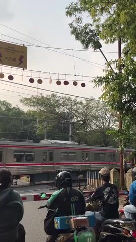 A train passes through a railroad crossing. People on motorcycles are waiting for the train to pass. The crossing has a yellow sign with a warning that reads "AWAS LISTRIK ALIRAN ATAS 4.7 M". There are decorative circles hanging on a wire above the crossing. Trees and foliage are visible in the background. The sky is cloudy.
