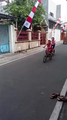 The video shows two young girls riding small bicycles down a paved street lined with houses and trees. Both girls are wearing casual clothing and appear to be of Asian descent. Indonesian flags are visible, suggesting the location may be Indonesia. The first girl, wearing a red shirt and shorts, is riding a dark-colored bicycle. The second girl, wearing a pink shirt and dark shorts, is riding a lighter-colored bicycle. A third girl, also in red, is seen further down the street on a bicycle, and a fourth girl, in pink, is visible even further in the distance. A motorcycle with a rider is passing by in the opposite direction. The street is mostly empty except for the children on their bikes, and the overall atmosphere seems peaceful and domestic. The lighting suggests it is daytime.