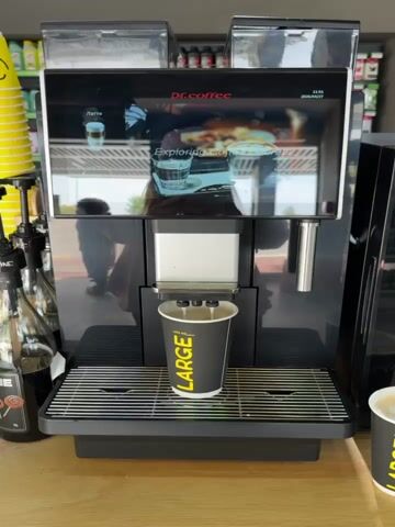 Here's a description of the video:

The video shows a coffee machine dispensing a beverage into a large paper cup. The machine has a screen that displays "dr.coffee" and an image of a coffee drink. Three nozzles are dispensing the liquid into the cup, which is labeled "LARGE" and "400 ml".  The machine is mostly black with stainless-steel accents. The background shows a person walking outside, visible through a window's reflection on the machine's screen. There are syrup bottles to the side of the machine.