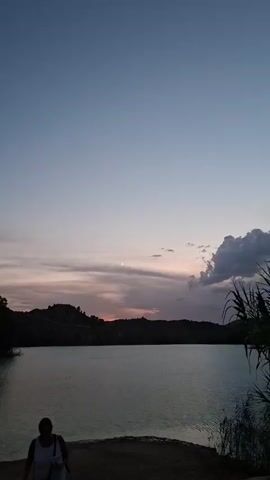 A serene twilight scene unfolds over a body of water. A sliver of a crescent moon hangs in the pale blue sky, partially obscured by soft, wispy clouds that catch the fading light of sunset, painting streaks of pink and orange across the horizon. Silhouetted against this dramatic sky are rolling hills in the distance and the dark, jagged outline of vegetation in the foreground on the right. In the lower left corner, a person stands partially visible, facing away from the camera, their form a dark silhouette against the illuminated water and sky. The water itself is calm, reflecting the muted colors of the sky, creating a tranquil and contemplative atmosphere.