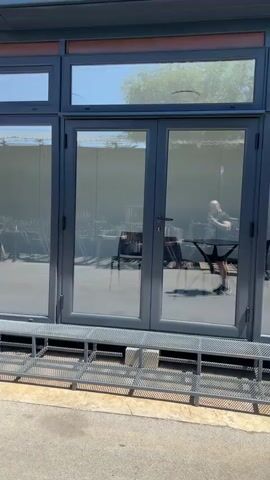 The video shows a person in a black t-shirt and black shorts standing in front of a glass-paneled door. The person is holding a phone and appears to be filming themselves or something within the building. The building has large windows and doors that reflect the surroundings. The environment appears to be outdoors, possibly a patio or a courtyard, with a concrete ground and a metal bench or seating area in front of the doors. The building's interior is partially visible through the glass, showing what looks like a kitchen or a workspace with cabinets and appliances. The person's reflection is prominent in the glass.