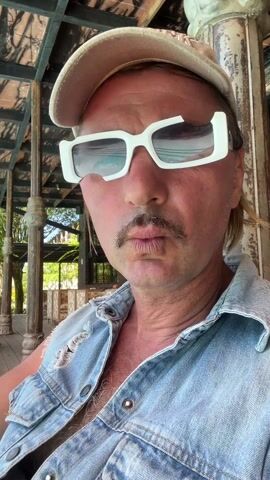 A man with a mustache, wearing a denim vest, a New York baseball cap, and white-framed sunglasses, is the main subject of the video. He's sitting under a covered structure, with wooden support beams visible. Behind him, there is a pool area with two women lounging on wooden chairs. One woman is using her phone, while the other is wearing a hat. The scene is set in a tropical environment with palm trees and other greenery visible in the background under a clear blue sky. The camera appears to be held by the man, who is making eye contact with the lens.