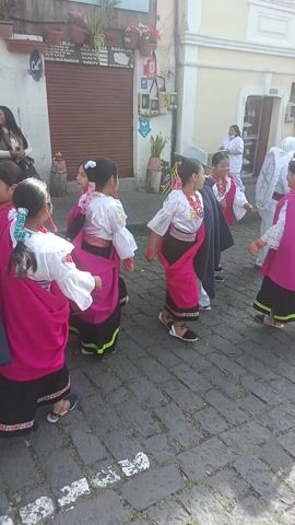 The video captures a group of young girls performing a traditional dance on a cobblestone street. The girls are dressed in matching traditional clothing, consisting of white embroidered blouses, black skirts with colorful trim, and bright pink shawls. They are also wearing beaded necklaces.  A young boy, dressed in a suit and fedora, appears to be leading the dance or acting as a partner. Several onlookers are standing on the side of the street, watching the performance. A building with a closed shutter is visible in the background, as are potted plants. The scene is well-lit, suggesting it is daytime.