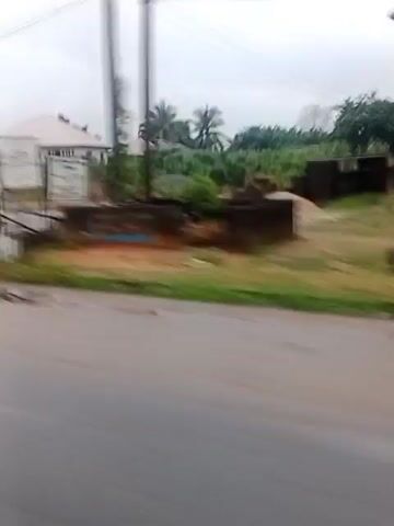 The video shows a blurry, moving shot of a street scene. A person in a white shirt and dark pants runs across a road. In the background, there are buildings under construction, some with multiple stories and balconies. There are also trees and vegetation visible. The sky appears overcast. The footage seems to be taken from a vehicle moving at a moderate speed.