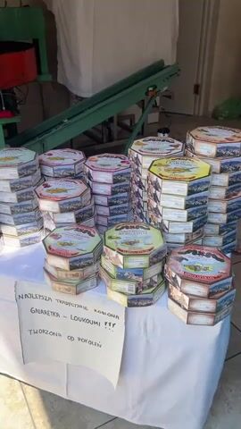 The video displays a factory setting with various pieces of machinery and stacked boxes of products. In the foreground, a table is covered with a white tablecloth, upon which are numerous stacks of octagonal boxes, likely containing sweets or food items. These boxes are colorful and feature images of landscapes and text in various languages. A handwritten sign is placed on the table, with text in Polish, indicating that the product is "NAJLEPSZA TRADYCYJNE KOBIENA GNARETKA - LOUKOUMI TWORZONA OD POKOLEŃ". To the left, a green industrial machine with various tubes, wires, and a red component is visible. Behind the table and machines, there are more stacks of boxes and a large piece of machinery covered with a white sheet. A conveyor belt, also green, runs from the front of the scene towards the back. The walls are a light beige color, and there is a doorway in the background with two white doors, one of which is slightly ajar. The lighting suggests an indoor environment, possibly a factory or workshop.