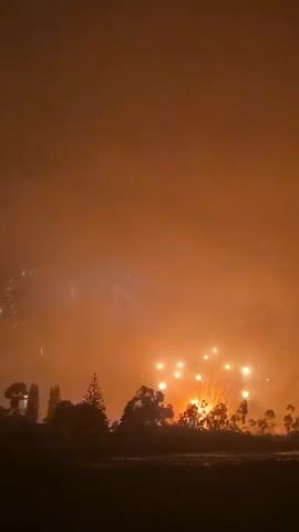 The video shows a fireworks display at night. In the foreground, a child's umbrella with Paw Patrol characters is visible. In the background, silhouetted trees and a body of water are seen. Fireworks are exploding in the sky, illuminating the scene with bright flashes of light and smoke. The colors of the fireworks range from yellow and orange to pink. The video pans slightly, showing the continuous bursts of fireworks.