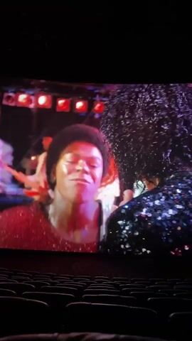 The video shows a movie screen in a darkened movie theater. The screen displays a scene of a concert featuring a young Michael Jackson. He is wearing a sparkly jacket and singing into a microphone, illuminated by stage lights. The camera angle looks up from the back rows of the theater, focusing on the screen. The seats in the theater are mostly empty.