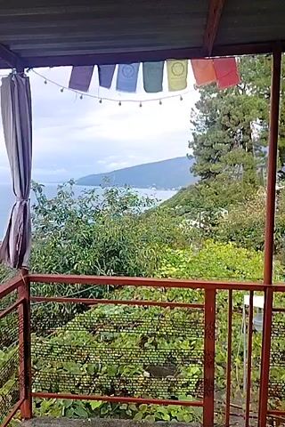 The video shows a view from a balcony or patio looking out at a coastline. The balcony has a corrugated metal roof and a rusted metal railing. Prayer flags hang from the roofline, and a curtain hangs to the left. Below the balcony is lush greenery, including what appears to be grapevines. In the distance, the sea stretches out to the horizon under a cloudy sky. A town or village is visible on the coast, with buildings and trees. The overall impression is one of a scenic, possibly tropical or Mediterranean, location.