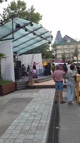 A live music event is taking place outdoors under a clear sky. A band is performing on a stage set up with speakers and instruments. A group of people are gathered, some standing and watching the performance, while others are seated at tables in the background. The environment appears to be a casual outdoor venue, possibly a bar or restaurant, with trees and buildings visible in the distance. The lighting suggests it is late afternoon or early evening.