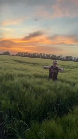 A man standing in a field of tall, green plants raises his arms into a Y. He is wearing a dark tank top with a Chicago logo on it and sunglasses. The field is lit by the orange glow of a sunset. The camera pulls back, showing the field in the distance. The man runs, ducking below the top of the plants and moving through the field.