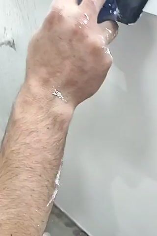 This video shows a person's hands applying a smoothing tool, possibly a drywall mud pan or a wide putty knife, to a white wall. The tool is metal with a black handle and is covered in white plaster or mud. The person's hands are also covered in the white substance. The wall appears to be under renovation or repair, as there are patches of missing paint and debris on the floor. The person is moving the tool horizontally across the wall, likely to smooth out a layer of joint compound. The video is shot from a close-up perspective, focusing on the action of plastering.