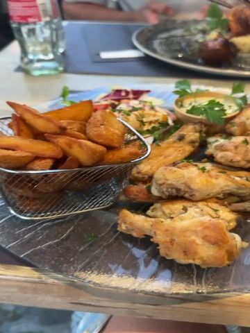 This video shows a close-up of a meal on a table, with two plates of food visible. The foreground plate contains chicken wings and potato wedges served in a metal basket, along with a small salad and a dipping sauce. The background plate features skewers of meat served with couscous, a piece of sweet potato, and a cherry tomato. A person's arm is visible in the foreground, and another person is partially visible in the background, holding a utensil. The meal appears to be in a restaurant setting, with other drinks and tableware also present.
