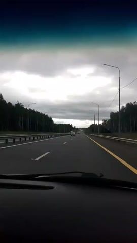 This media file captures a first-person perspective from inside a vehicle driving on a highway. The scene is set on a multi-lane road with guardrails and streetlights lining both sides. Dense forest borders the highway, suggesting a rural or suburban setting, consistent with the specified location of Larionovo, Russia.

The sky is heavily overcast with dark, dramatic clouds, indicating a gloomy or potentially stormy weather condition. The lighting is subdued, characteristic of a cloudy day. Several vehicles are visible on the road, including cars and a large truck with a distinctive orange trailer. The truck has visible text on its side, reading "Delko" in white lettering.

The activity depicted is straightforward travel on a highway. The overall mood is somewhat somber due to the weather, but the presence of other vehicles suggests ongoing activity and movement. There are no people explicitly visible, but their presence is implied by the vehicles. The time of day is not definitively clear but appears to be daytime, given the visibility and the activity on the road.