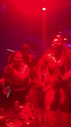 The video shows a dimly lit nightclub with red and purple lighting. Several people are dancing and socializing. In the foreground, two women are dancing close together. A bottle of alcohol and several shot glasses are visible on a table. The overall atmosphere is energetic and festive.