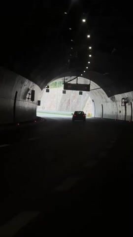 The video shows a car driving through a tunnel. The tunnel is illuminated by lights overhead, and the car is approaching the exit. Another car is visible ahead, already exiting the tunnel. The surroundings outside the tunnel consist of a road, a rock face, and sparse vegetation. The scene is captured from a static viewpoint within the tunnel, focusing on the cars and the approaching exit. A sign indicating a speed limit of 100 is hanging from the tunnel's ceiling.