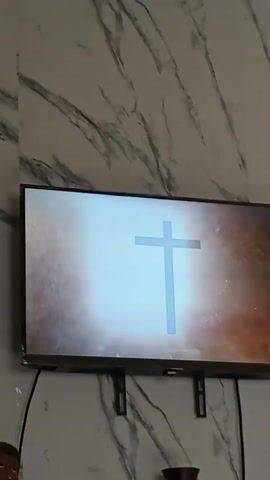 A television mounted on a wall is displaying a graphic. The graphic begins with a black cross against a background of clouds illuminated by light. The cross gradually transforms into the word "JESÚS." The wall behind the television has a marble-like pattern. A small, dark object is visible below the television.