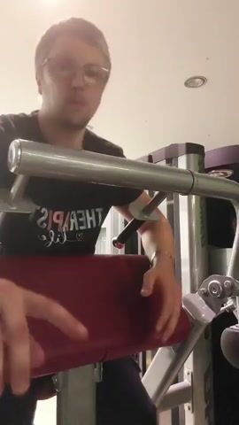 A person with glasses and blonde hair is performing bicep curls on a weight machine in a gym. They are wearing a black t-shirt with white text and black pants. The machine has a red padded seat and silver metal bars. The person is gripping the handles of the machine with both hands and is in the process of curling the weights upwards. The background shows gym equipment and lighting fixtures.