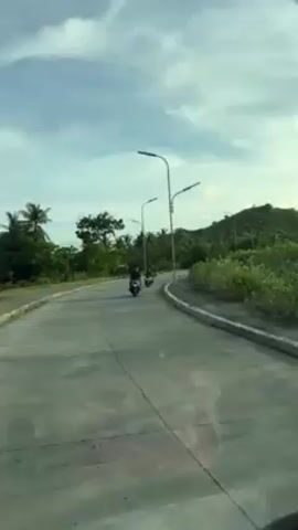The video shows a road with a motorcycle and a car on it. The road is paved and has street lights along the side. There are trees and a hill in the background. The sky is cloudy. The camera appears to be inside a moving vehicle. There is also audio in the video of people speaking.