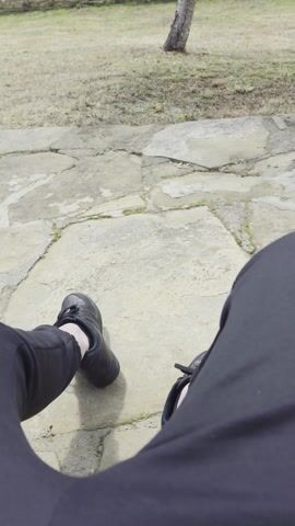 The video shows a first-person view of someone sitting outside on a stone patio. The person is wearing black shoes and black pants. In the background, there is a building with a stone base and a light yellow-green upper section. A tree with white blossoms is in full bloom in the foreground, slightly obscuring the view of the building. The grass is dry and brown, and there are patches of green. The sky is visible through the tree branches, appearing bright and overcast. Another smaller building with a stone base and slate roof can be seen to the right. The camera slowly pans up, focusing on the blossoming tree and the architecture of the buildings.