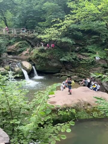The video begins with a view of a waterfall cascading into a pool surrounded by lush green trees. A wooden bridge crosses over a stream in the background. Several people are visible on the bridge and on a path near the waterfall. The camera then focuses on a person wearing a backpack, standing on a rock ledge. The person turns and walks along the ledge, revealing more of the surrounding forest and a road. Other people are seen walking along the road and sitting on a bench. The video pans to show a group of people gathered near the waterfall, some of whom appear to be taking pictures. The scene is set in a natural, outdoor environment with a focus on the waterfall and the surrounding vegetation.