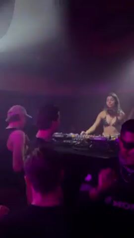 The video shows a DJ playing music at a club. The DJ is a woman wearing a bikini top. She is standing behind a mixing console. There are people in the audience dancing. The club is dark and the lights are purple. One man in the audience wears sunglasses and a Honda-branded top.