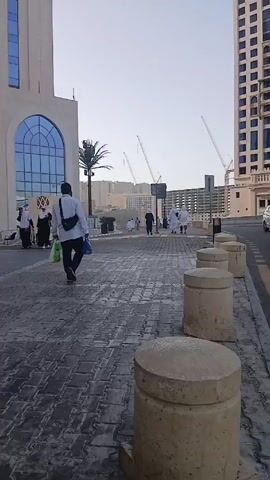 The camera records a static shot of a street scene. A sidewalk paved with dark gray bricks runs along the left side of the frame, separated from the road by a series of short, cylindrical concrete barriers. People, dressed in both traditional white clothing and casual attire, are walking in the distance on the sidewalk. 

To the right of the sidewalk is a two-lane road. A black SUV drives along the road. On the other side of the road, a tall building with a beige facade and many windows dominates the background. Several cranes are visible in the distance, suggesting ongoing construction. A pale sky provides a neutral backdrop to the urban environment.
