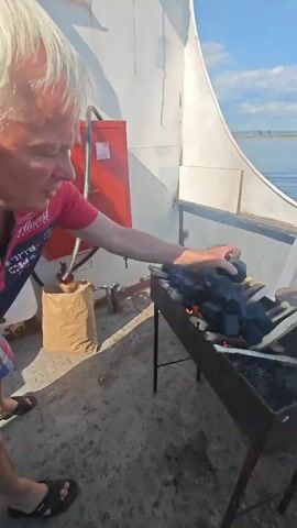 A man wearing a pink shirt and blue apron is tending to a charcoal grill on the deck of a boat. He is reaching towards the grill, which is filled with charcoal and wood, and appears to be in the process of lighting it, as evidenced by the smoke and flames. The boat is on the water, with a shoreline visible in the distance. The deck is concrete, and there is a brown paper bag and a red fire extinguisher on the left side of the frame. The sky is blue with some clouds.